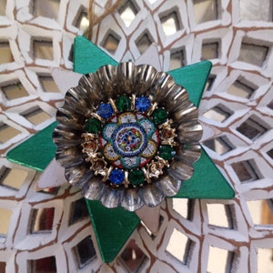 May include: A silver and gold star-shaped ornament with a mosaic design in the center. The mosaic features blue, green, and yellow glass pieces. The ornament is hanging from a white string.