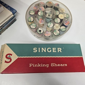 May include: A clear plastic container filled with colorful spools of thread sits above a box of Singer pinking shears. The box is teal and red with the Singer logo and text.