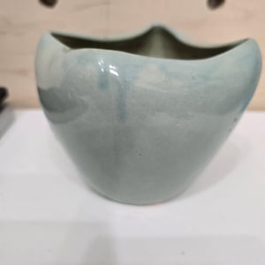 May include: A light blue ceramic vase with a unique, wavy rim. The vase has a smooth, glossy finish and a rounded, asymmetrical shape. The interior is a darker shade of blue-green. This decorative vase is suitable for displaying flowers or as a standalone art piece.