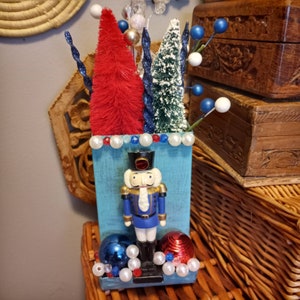 May include: A blue painted wooden box with a nutcracker figurine, red and white Christmas trees, and white and blue beads.