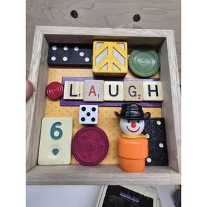 May include: A wooden framed collage with the word "LAUGH" spelled out using Scrabble tiles. The collage includes a domino, a peace sign, a dice, a clown figure, and other game pieces. The background is yellow.