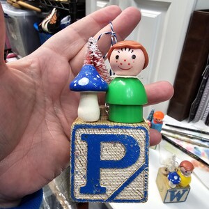 May include: A wooden block ornament with the letter "P" painted in blue. A small plastic figure with a green shirt and a red hat stands on top of the block. A blue and white mushroom with a white stem is attached to the figure's side. A small red and white brush is attached to the top of the figure's head.