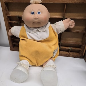 May include: A doll with a round head, blue eyes, and a small tuft of blonde hair on top. The doll wears a yellow corduroy romper over a white collared shirt with two buttons, white socks, and white shoes. A small elephant patch is on the romper.