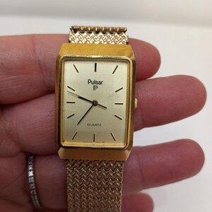 May include: A gold-toned Pulsar watch with a rectangular face and a gold mesh band. The watch face has a white background with black hour and minute hands, black Roman numeral hour markers, and the words "Pulsar P" and "Quartz" printed in black.