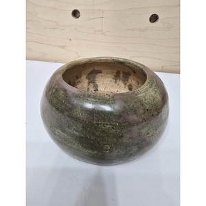 May include: A round, ceramic bowl with a wide opening. The bowl has a mottled green and purple exterior with a lighter interior. The handmade pottery has a rustic, organic aesthetic, suitable for home decor or as a planter.