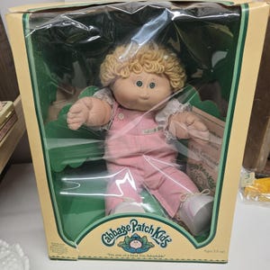 May include: A Cabbage Patch Kids doll with blonde yarn hair and green eyes, wearing pink overalls and a white shirt. The doll is inside a yellow and green cardboard box with a clear plastic window, featuring the text Cabbage Patch Kids.