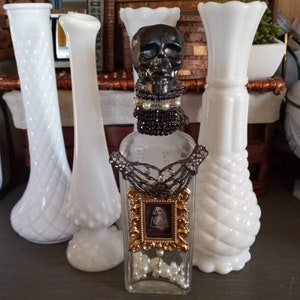 May include: A clear glass bottle with a skull bottle stopper, black beads, and a gold picture frame with a skeleton inside. The bottle is surrounded by three white milk glass vases.