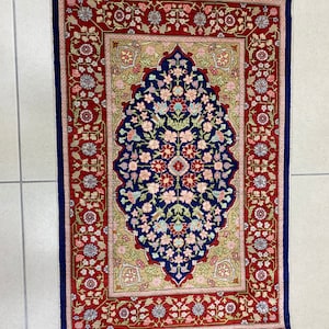May include: A rectangular rug with a central floral design in navy blue, surrounded by a beige field and a red border. The border features floral patterns and a navy blue edge with fringe.
