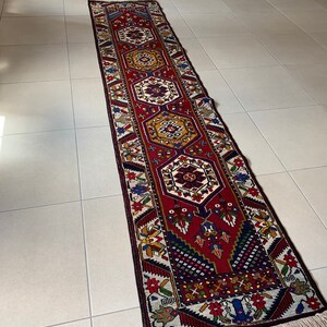 Op de afbeelding: Een lange, rechthoekige loper met een gedetailleerd geometrisch patroon. De loper heeft een rood middenveld met zich herhalende medaillons in tinten goud, blauw en wit. De rand is donker met bloemen- en geometrische motieven en de loper heeft een gefranjerde rand.