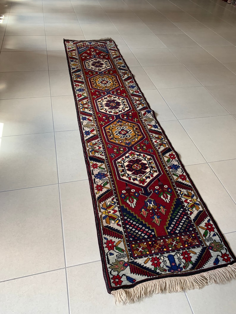 Op de afbeelding: Een lange, rechthoekige loper met een gedetailleerd geometrisch patroon. De loper heeft een rood middenveld met zich herhalende medaillons in tinten goud, blauw en wit. De rand is donker met bloemen- en geometrische motieven en de loper heeft een gefranjerde rand.