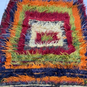 Könnte beinhalten: Ein farbenfroher, rechteckiger Teppich mit zotteliger Textur. Der Teppich zeigt ein zentrales quadratisches Design in Rot und Weiß, umgeben von konzentrischen Bändern in Orange, Grün und Blau. Die Kanten haben einen Fransen.