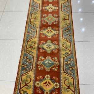 Puede incluir: Una alfombra de pasillo rectangular con un patrón intrincado. La alfombra presenta una banda central de color marrón rojizo con motivos repetidos de diamantes y flores en tonos de amarillo, azul y rosa. Los bordes son una mezcla de amarillo y azul, creando un diseño simétrico.