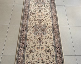 Op de afbeelding: Een rechthoekig beige loperkleed met een ingewikkeld bloemenpatroon in bruin-, grijs- en crèmetinten. Het kleed heeft een gedetailleerde rand en franjes aan beide uiteinden.