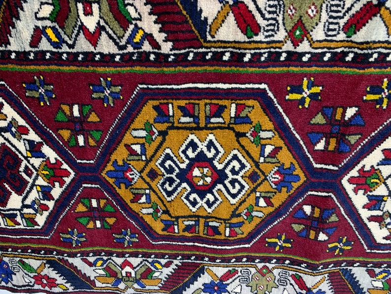Op de afbeelding: Close-up van een gedetailleerd, patroonrijk tapijt met een rijk kleurenpalet. Het ontwerp bevat geometrische vormen, waaronder een centrale zeshoek en ruitvormen, in tinten bordeaux, goud, blauw en cr&egrave;me. Het tapijt heeft een traditionele, sierlijke stijl.