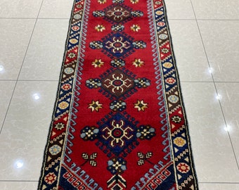 Op de afbeelding: Een rechthoekig rood loperkleed met een geometrisch patroon. Het kleed heeft een centraal rood veld met blauwe, bruine en crèmekleurige geometrische patronen. De rand is donkerblauw met herhalende patronen. Het kleed is waarschijnlijk van wol.