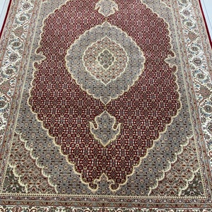 May include: A large, rectangular area rug with an intricate, symmetrical pattern. The rug features a deep red central field with a repeating geometric design. A large, ornate medallion in shades of gray and beige dominates the center. The border is cream-colored with floral motifs.