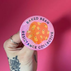 May include: A round, pink button with the text "BAKED BEAN RESISTANCE COLLECTIVE" in a circular pattern. The center features an orange and yellow graphic of baked beans. The button has a glittery, iridescent finish.