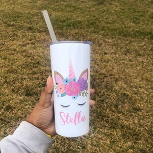 Peut inclure: Un gobelet en acier inoxydable blanc avec un motif de licorne orn&eacute; de fleurs roses, violettes et bleues. Le gobelet est dot&eacute; d'une paille et le nom "Stella" est imprim&eacute; sur le devant.