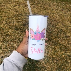 Peut inclure: Un gobelet en acier inoxydable blanc avec un motif de licorne et le nom "Stella" imprim&eacute; dessus. La licorne a une corne rose, des oreilles roses et une couronne florale avec des fleurs roses, violettes et bleues.