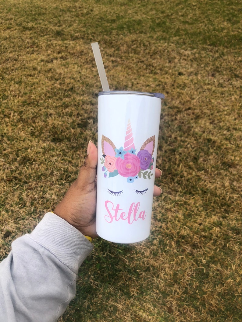 Peut inclure: Un gobelet en acier inoxydable blanc avec un motif de licorne et le nom "Stella" imprim&eacute; dessus. La licorne a une corne rose, des oreilles roses et une couronne florale avec des fleurs roses, violettes et bleues.