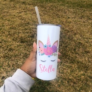 Peut inclure: Un gobelet en acier inoxydable blanc avec un motif de licorne orn&eacute; de fleurs roses et violettes, des yeux ferm&eacute;s et une corne dor&eacute;e. Le gobelet porte le nom "Stella" en lettres roses.