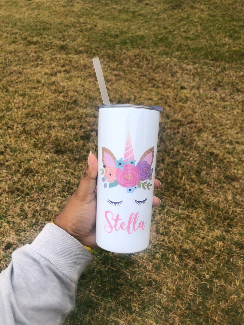 Peut inclure: Un gobelet en acier inoxydable blanc avec un motif de licorne orn&eacute; de fleurs roses et violettes, des yeux ferm&eacute;s et une corne dor&eacute;e. Le gobelet porte le nom "Stella" en lettres roses.