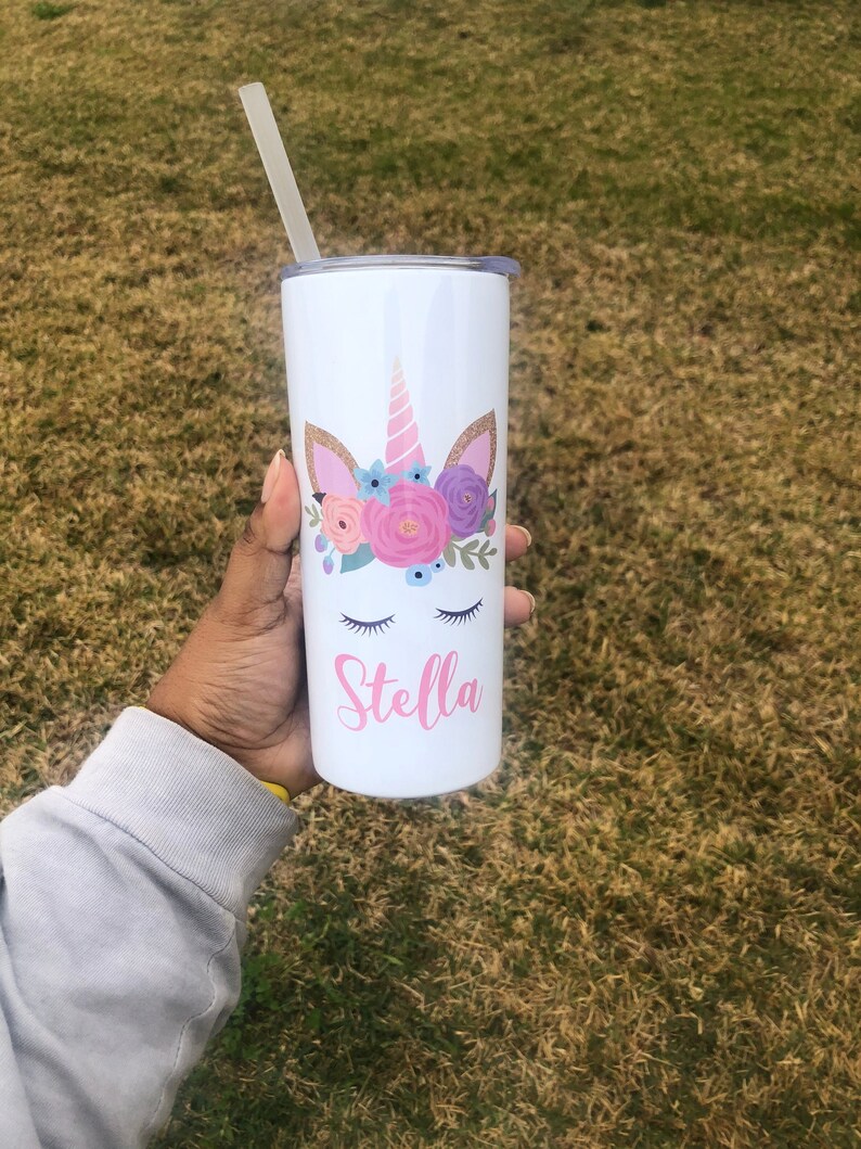 Peut inclure: Un gobelet en acier inoxydable blanc avec un motif de licorne orn&eacute; de fleurs roses, violettes et bleues. Le gobelet est dot&eacute; d'une paille et le nom "Stella" est imprim&eacute; sur le devant.
