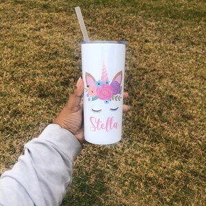 Peut inclure: Un gobelet en acier inoxydable blanc avec un motif de licorne et des fleurs roses. Le gobelet est muni d'une paille et le nom "Stella" est inscrit sur le devant.