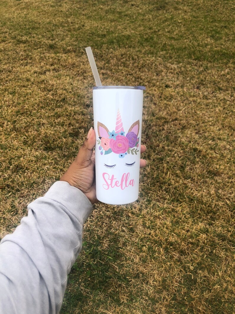 Peut inclure: Un gobelet en acier inoxydable blanc avec un motif de licorne et des fleurs roses. Le gobelet est muni d'une paille et le nom "Stella" est inscrit sur le devant.