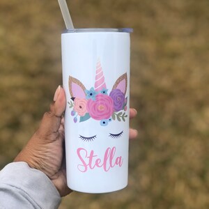 Peut inclure: Un gobelet en acier inoxydable blanc avec un motif de licorne orn&eacute; de fleurs roses, violettes et bleues, d'accents de paillettes dor&eacute;es et d'yeux ferm&eacute;s. Le nom "Stella" est &eacute;crit en rose sous la licorne.