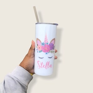 Peut inclure: Un gobelet en acier inoxydable blanc avec un motif de licorne orn&eacute; de fleurs roses, violettes et bleues. La licorne a une corne rose et des oreilles paillet&eacute;es dor&eacute;es. Le nom "Stella" est &eacute;crit en rose sous la licorne.