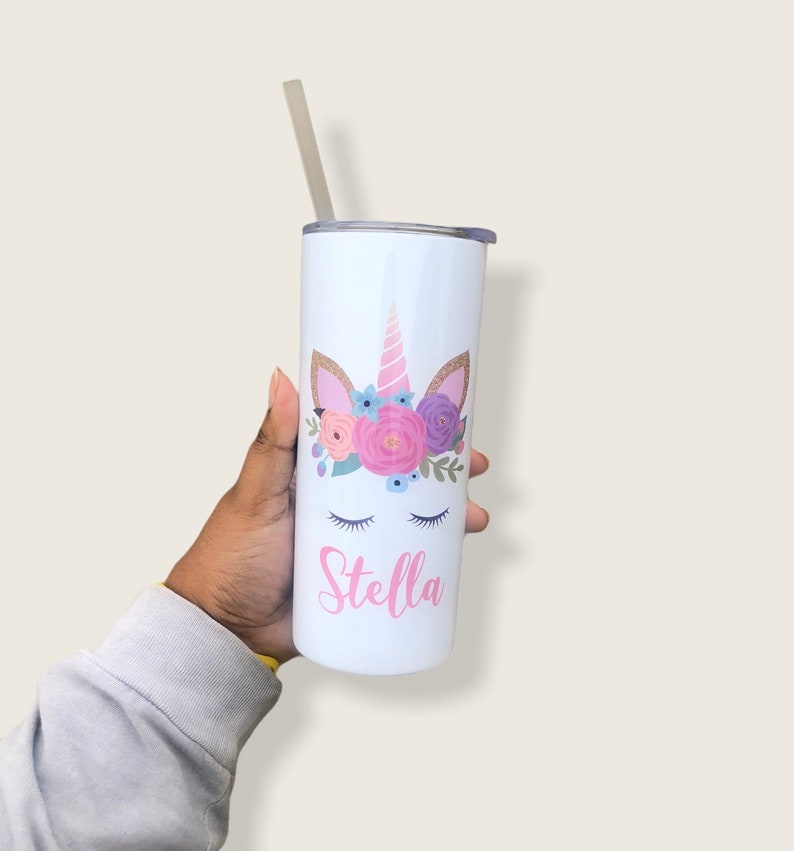 Peut inclure: Un gobelet en acier inoxydable blanc avec un motif de licorne orn&eacute; de fleurs roses, violettes et bleues. La licorne a une corne rose et des oreilles paillet&eacute;es dor&eacute;es. Le nom "Stella" est &eacute;crit en rose sous la licorne.