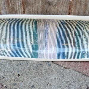 May include: An elongated, oval-shaped ceramic tray with a smooth, cream-colored rim. The interior surface features a repeating pattern of vertical stripes in shades of teal, navy blue, and soft lavender, with a glossy, reflective finish throughout.