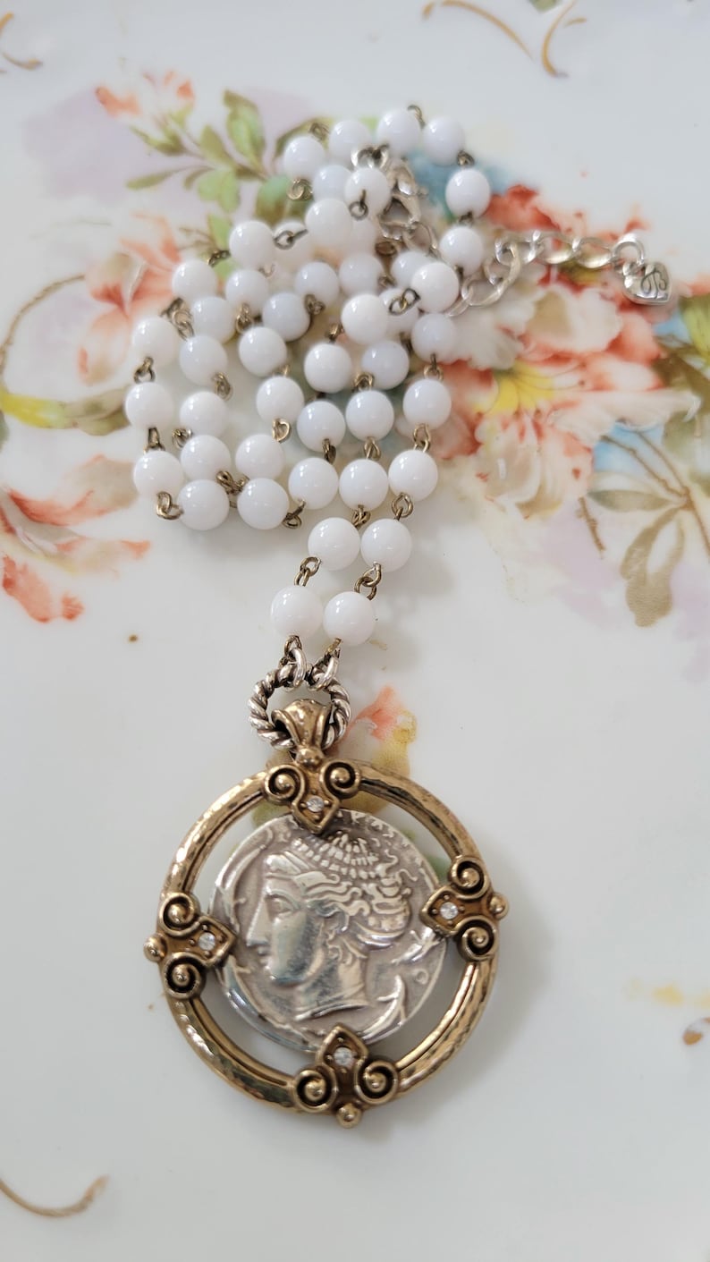 May include: A vintage necklace featuring a silver coin pendant with a profile of a woman, set in a gold-toned frame. The necklace has a strand of white, round beads and a silver chain.