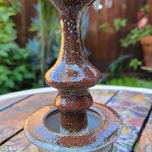 May include: A handcrafted ceramic candle holder with a speckled brown and white glaze. The design features a wide base, a central column, and a flared top. The piece is suitable for a standard-sized candle.