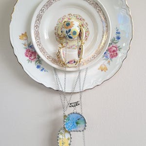May include: A decorative wall hanging featuring vintage floral plates and a teacup. Three necklaces with floral designs hang below. The plates have gold trim and pink, blue, and yellow floral patterns. The necklaces have silver chains.