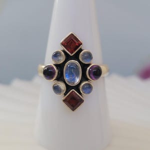 May include: A silver ring with an ornate design featuring an oval blue gemstone at the center, surrounded by smaller round and square gemstones in shades of red, blue, and purple. The ring is displayed on a white cone.