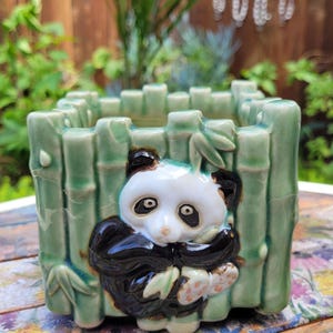 May include: A square ceramic planter with a panda bear design. The planter is light green with bamboo-style detailing. The panda has a white face, black ears, and black and white body. The planter is sitting on a floral patterned surface.