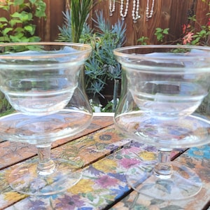 May include: Two clear glass goblets with a unique double-bowl design. The upper bowl is smaller and sits inside the larger lower bowl. The rims have a gold trim. The glasses are on a floral patterned table, with greenery in the background.