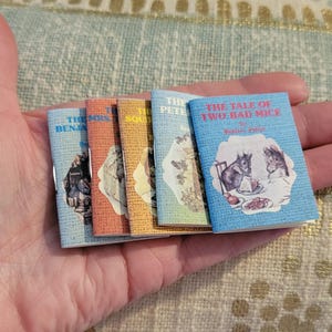 May include: A set of six miniature hardcover books with colorful covers. The titles include "The Tale of Two Bad Mice" by Beatrix Potter, "The Mrs. Tiggy-Winkle" by Beatrix Potter, "The Squirrel Nutkin" by Beatrix Potter, "The Peter Rabbit" by Beatrix Potter, and two others.