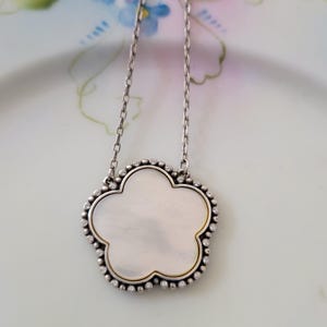 May include: Silver-tone necklace with a flower-shaped pendant. The pendant has a mother-of-pearl center, a gold-tone border, and a beaded silver-tone edge. The necklace is displayed on a floral patterned plate.