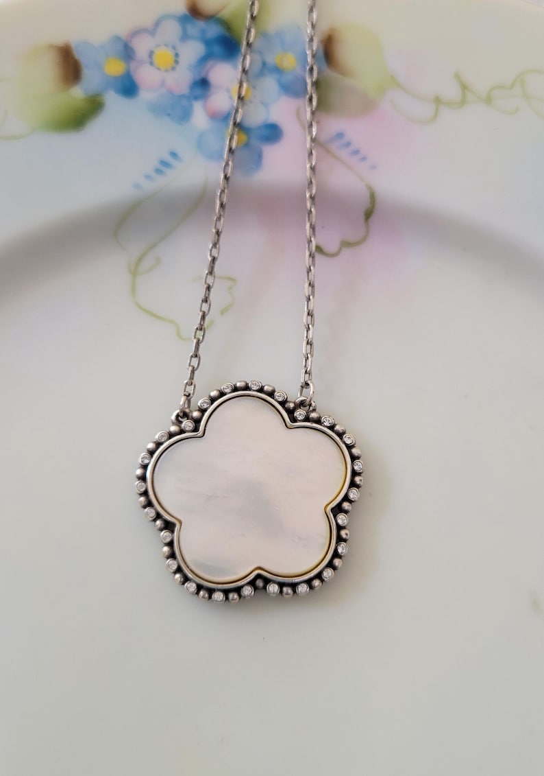 May include: Silver-tone necklace with a flower-shaped pendant. The pendant has a mother-of-pearl center, a gold-tone border, and a beaded silver-tone edge. The necklace is displayed on a floral patterned plate.
