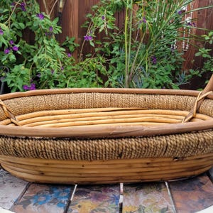 May include: An oval-shaped basket constructed from light brown rattan and natural fiber rope. The basket features a woven base, a reinforced rim, and two sturdy, curved handles attached to the sides. The design incorporates a textured rope band.