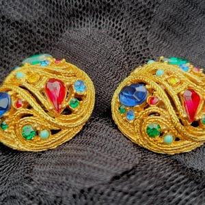 May include: A pair of gold-toned clip-on earrings. Each earring features a woven design and is adorned with colorful gemstones, including red, blue, green, and yellow. The earrings are set against a dark, textured background.