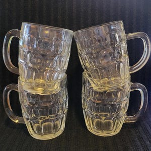 May include: Four clear glass beer mugs with a textured, dimpled design. Each mug has a large, curved handle. The mugs are stacked in two pairs, creating a symmetrical arrangement. The glass appears to be thick and durable.
