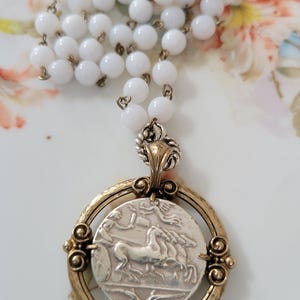 May include: A vintage-style necklace featuring a silver pendant with a detailed image of a horse-drawn chariot within a gold-toned frame. The necklace has a strand of white, round beads and a silver chain.