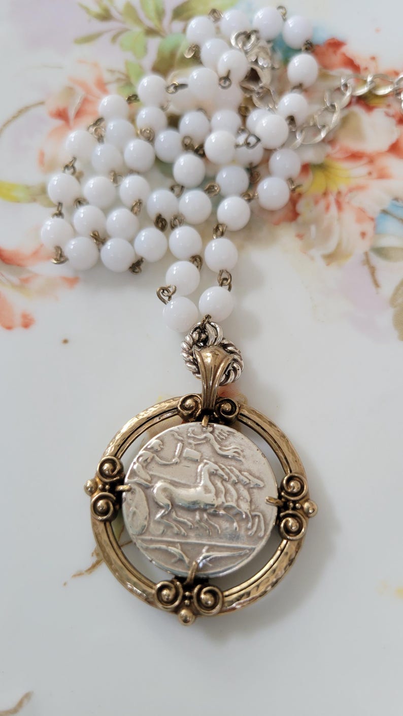 May include: A vintage-style necklace featuring a silver pendant with a detailed image of a horse-drawn chariot within a gold-toned frame. The necklace has a strand of white, round beads and a silver chain.