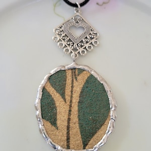 May include: A silver pendant necklace with a black cord. The pendant features a silver filigree design with a heart-shaped cutout above a large, oval-shaped pendant with a green and tan design.