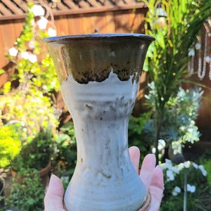 May include: A ceramic goblet with a flared rim and a tapered waist. The upper portion features a dark brown glaze, while the lower body is finished in a textured, off-white glaze. The base is circular and reveals the raw, reddish-brown clay.
