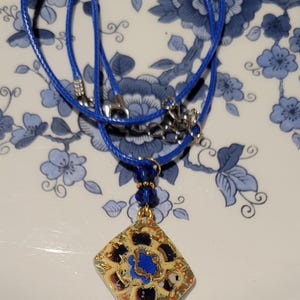 May include: A blue cord necklace with a square pendant featuring a colorful floral design with gold accents.